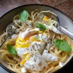 Spaghetti in a white bowl with a cracked sunny side up egg and basil leaves.