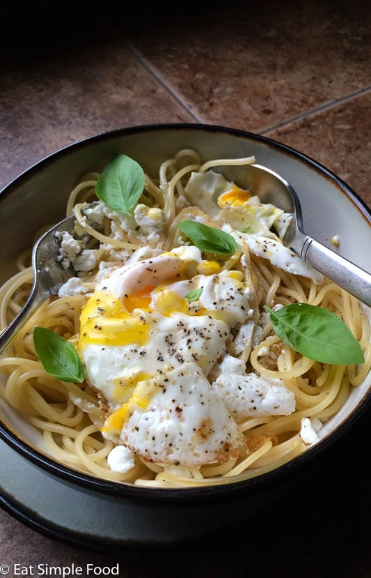 Spaghetti in a white bowl with a cracked sunny side up egg and basil leaves. 