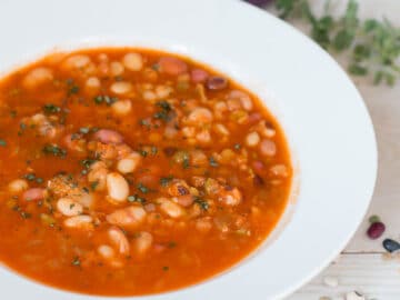 Side View of 13 bean soup in a red tomato sauce on a white wood table. side view. white bowl. Thyme sprigs in the back ground