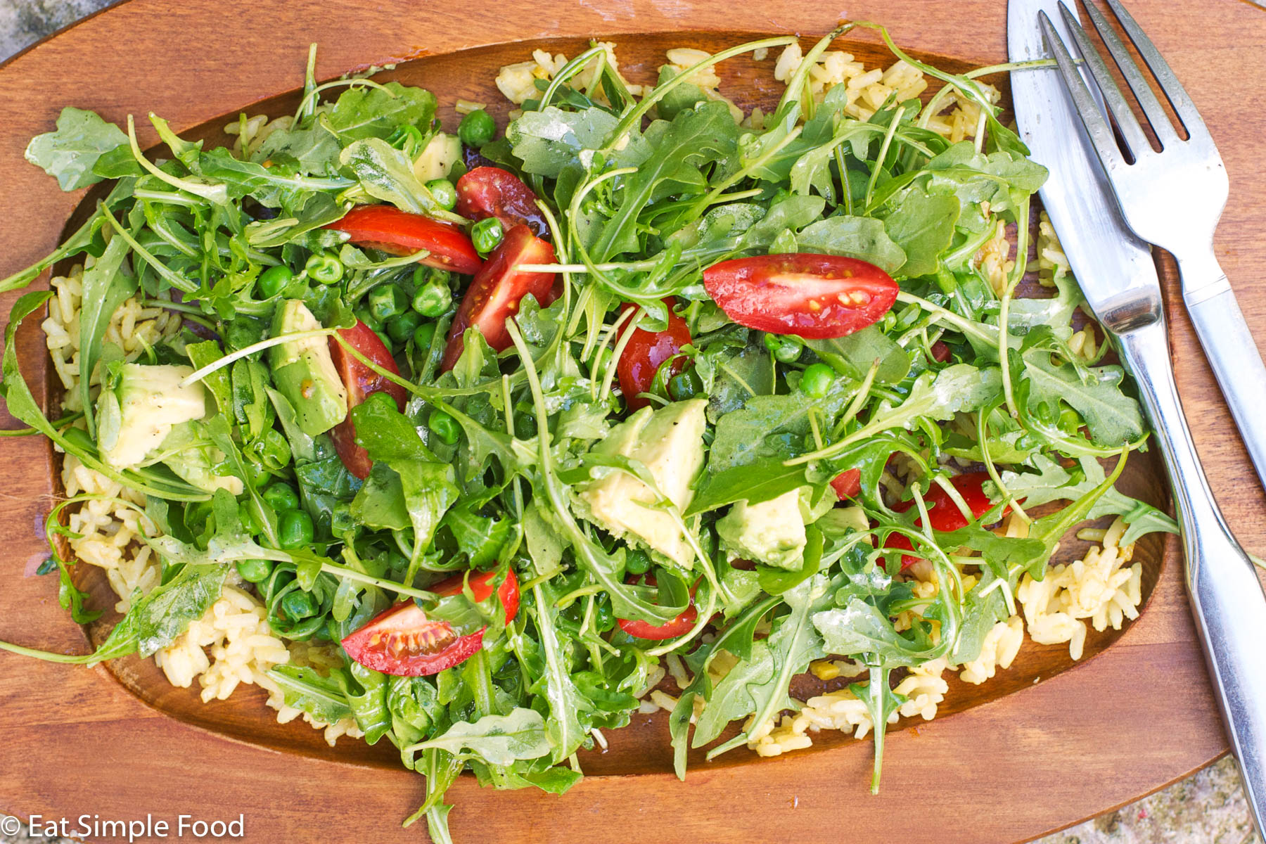 Brown oval plate of layered yellow rice, arugula, quartered cherry tomatoes, fresh spring peas, and chunks of avocado.