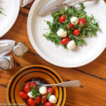 Arugula Salad topped with small mozzarella balls and cherry tomatoes on two white plates with knife and fork and salt and pepper shakers. top view.