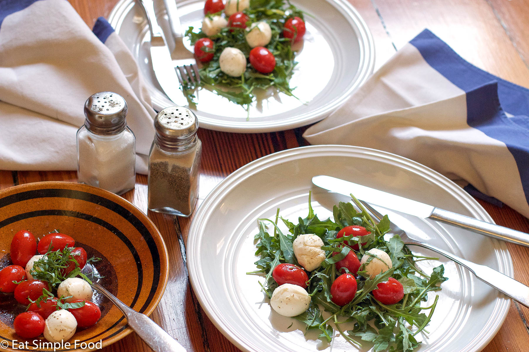 Arugula Salad topped with small mozzarella balls and cherry tomatoes on two white plates with knife and fork and salt and pepper shakers. side view.