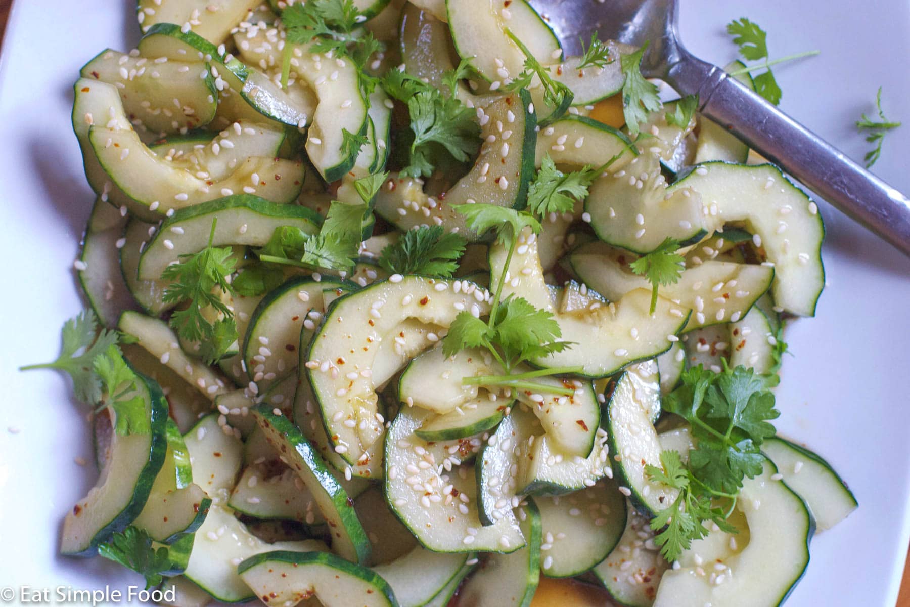 Small white plate of halved cucumbers with seeds taken out tossed in a soy / sesame oil sauce and topped with sesame seeds and cilantro.