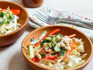 2 wood bowls of cole slaw with sliced red peppers, green onions and celery with cilantro and peanut garnish. Dressing, two forks and napkins in background.