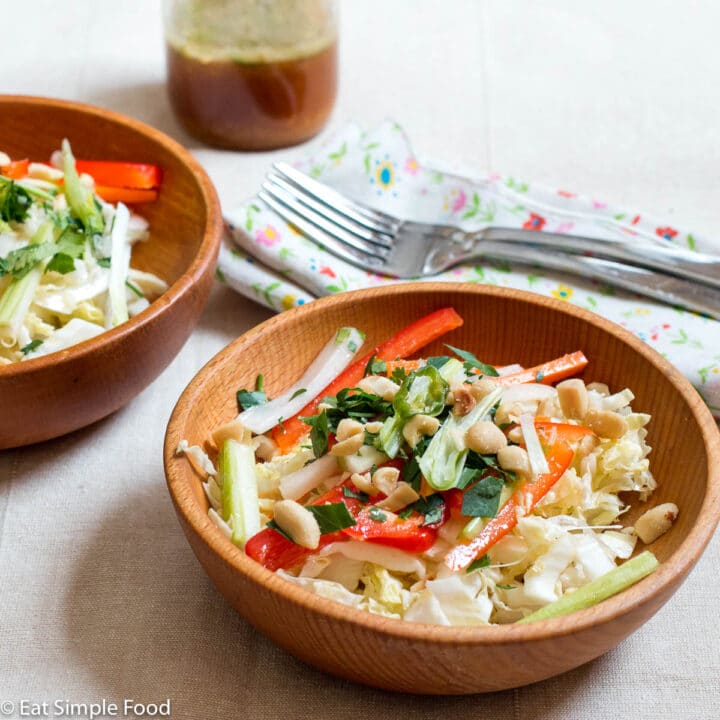2 wood bowls of cole slaw with sliced red peppers, green onions and celery with cilantro and peanut garnish. Dressing, two forks and napkins in background.
