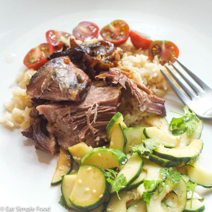 Close up of Pork Shoulder chunked and shredded over rice with halved cherry tomatoes and Cucumber Salad w/ Cilantro and Sesame Seeds.