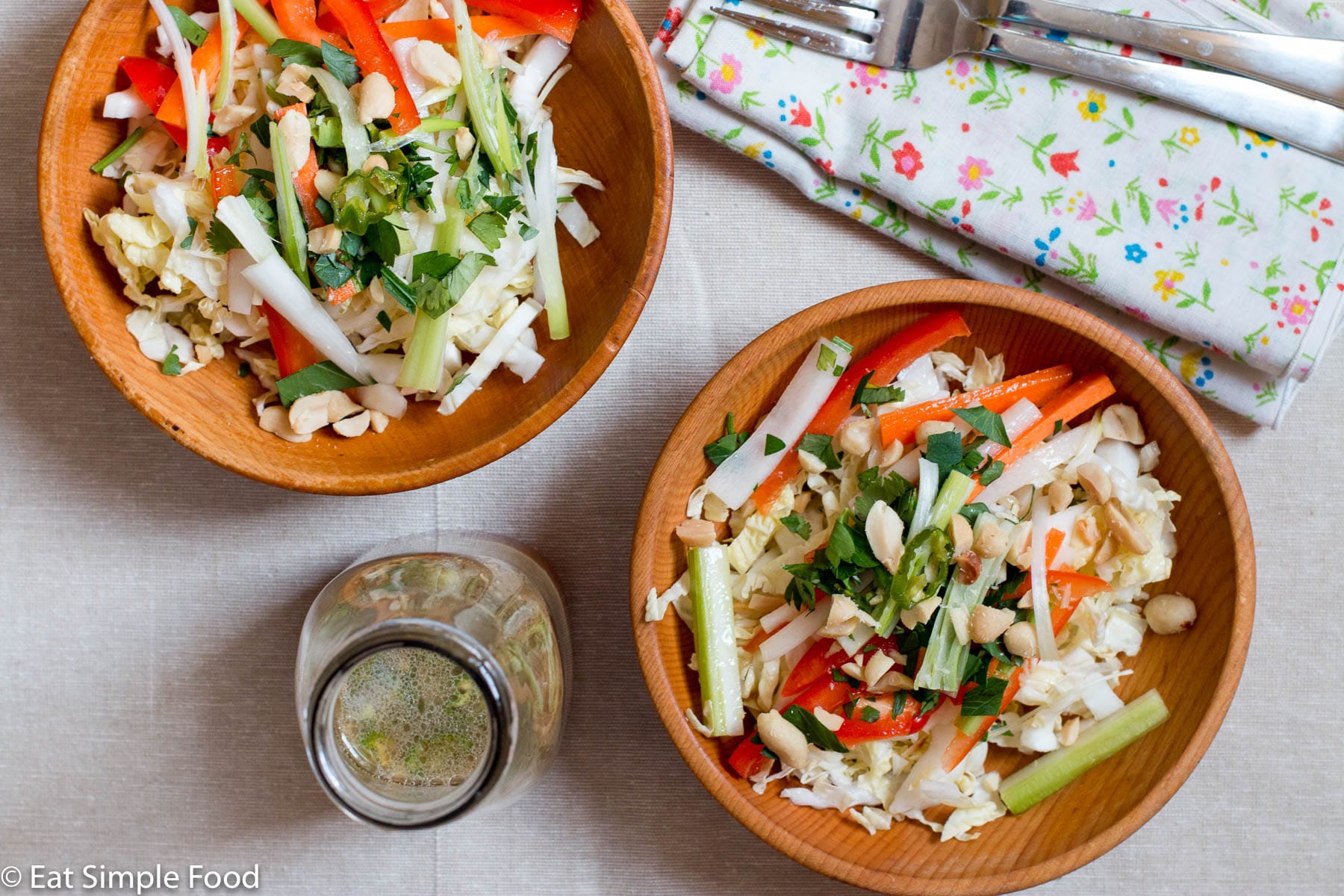 2 wood bowls of cole slaw with sliced red peppers, green onions and celery with cilantro and peanut garnish. Dressing, two forks and napkins in background. Top view.