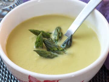 Small white bowl of creamy asparagus soup with asparagus tip garnish sticking out the top. Silver spoon in bowl. side view.