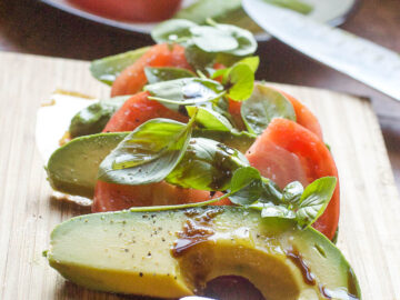 Sliced avocado, sliced tomato, basil leaves all layered on a wood cutting board with a balsamic sauce drizzled on top.