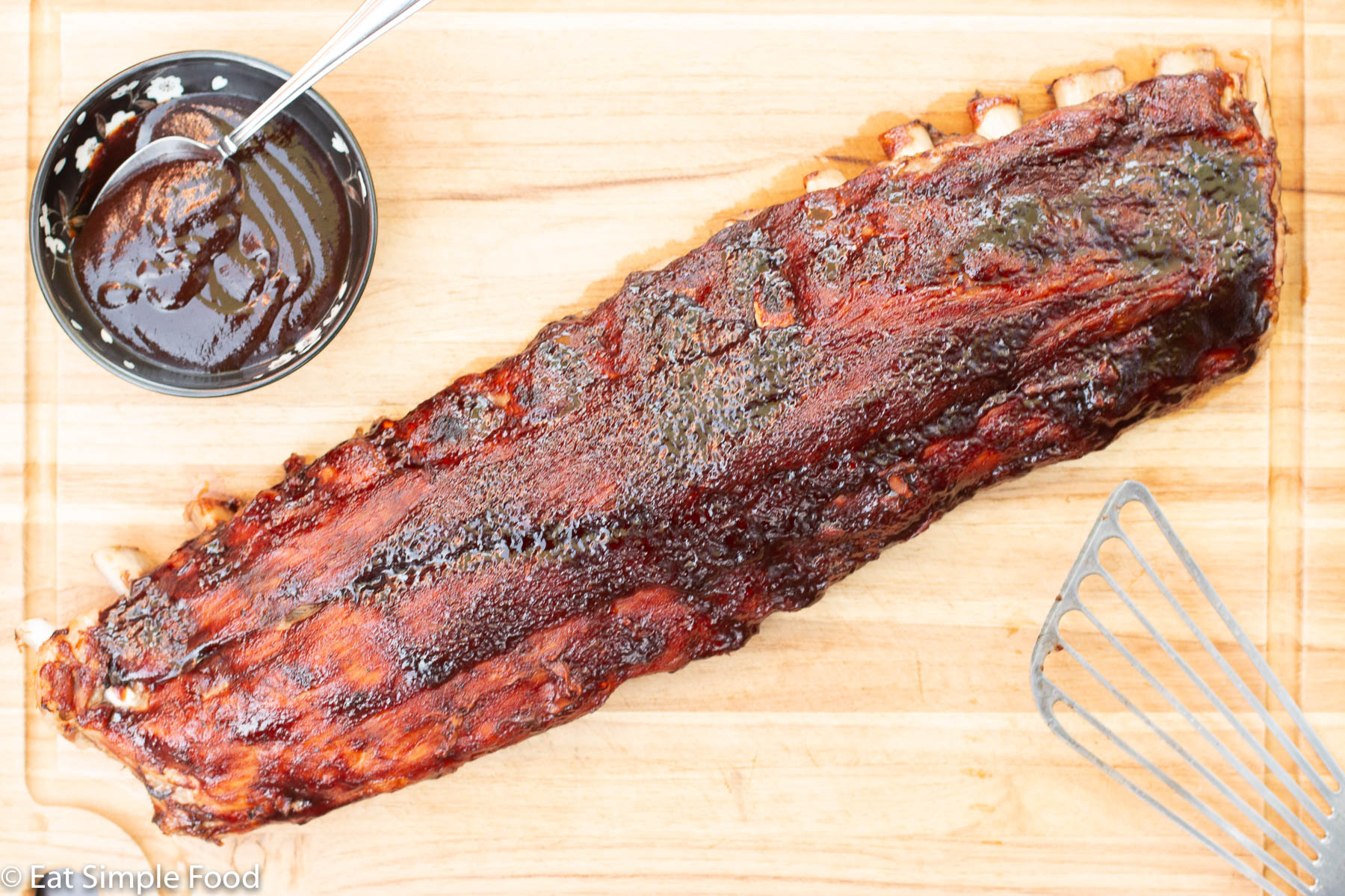 Whole Cooked Baby Back Ribs With BBQ Sauce Broiled On with a Metal Spatula and small black bowl of bbq sauce on a wood cutting board. Top view.