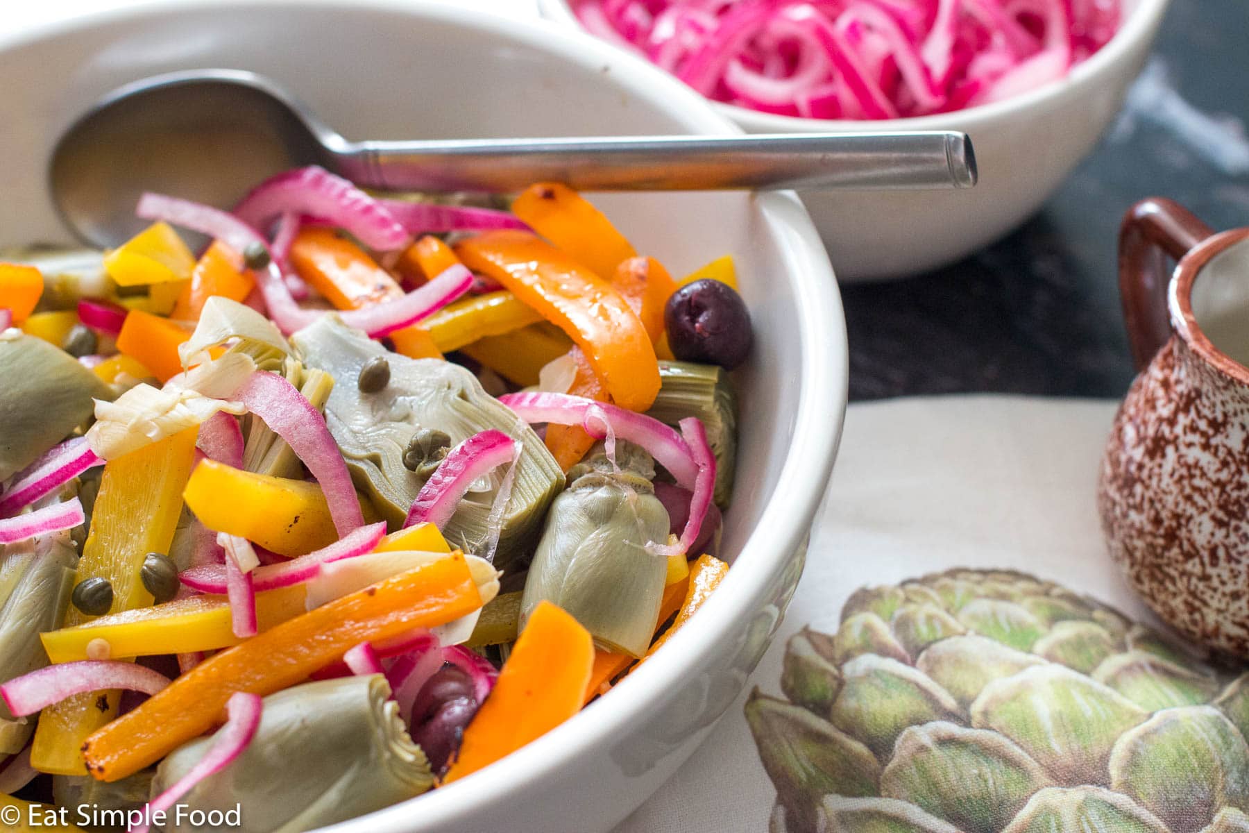 Side view of artichoke salad with kalamata olives, sliced orange peppers, capers and a bowl of pickled red onions on the side.