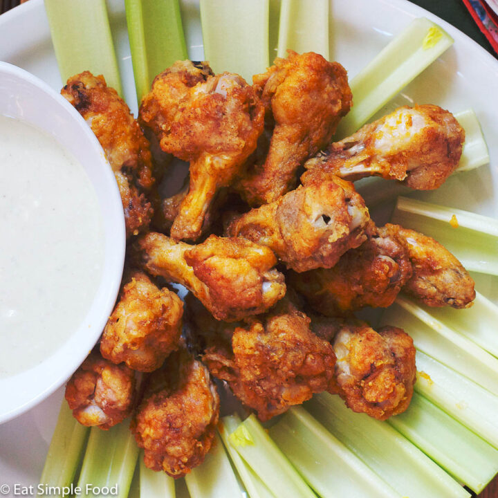 Spicy Red/Brown Chicken Drumsticks On A White Plate surrounded by celery and with with a white blue cheese dipping sauce in a bowl