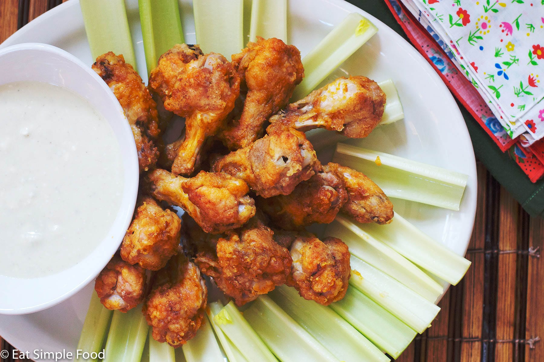 Spicy Red/Brown Chicken Drumsticks On A White Plate surrounded by celery and with with a white blue cheese dipping sauce in a bowl