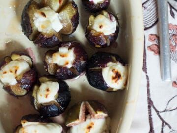 Figs with an x cut in the top and baked and stuffed with Goat cheese and drizzled with honey. Oval off white baking dish.