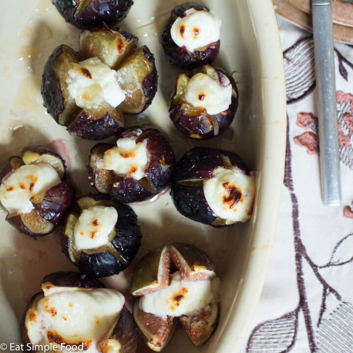 Figs with an x cut in the top and baked and stuffed with Goat cheese and drizzled with honey. Oval off white baking dish.