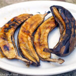 3 Grilled Banana Halves and One Whole, still in skin & grilled. Set on a white plate and has a honey butter glaze.