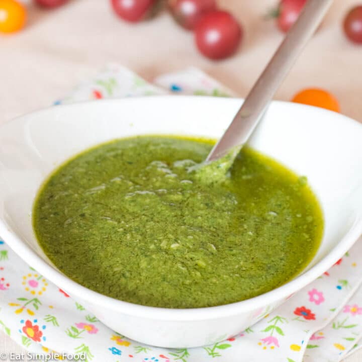Close up. Fresh Creamy basil pesto in a small white bowl on a delicate flowered napkin. Spoon sticking out and cherry tomatoes in background.