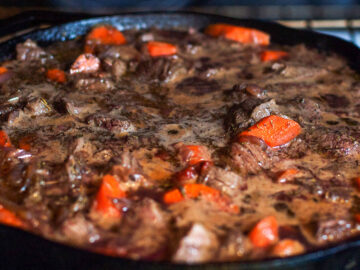 A Cast Iron Skillet over a gas oven cooking with chunked beef and carrots in a brown dark red wine gravy.