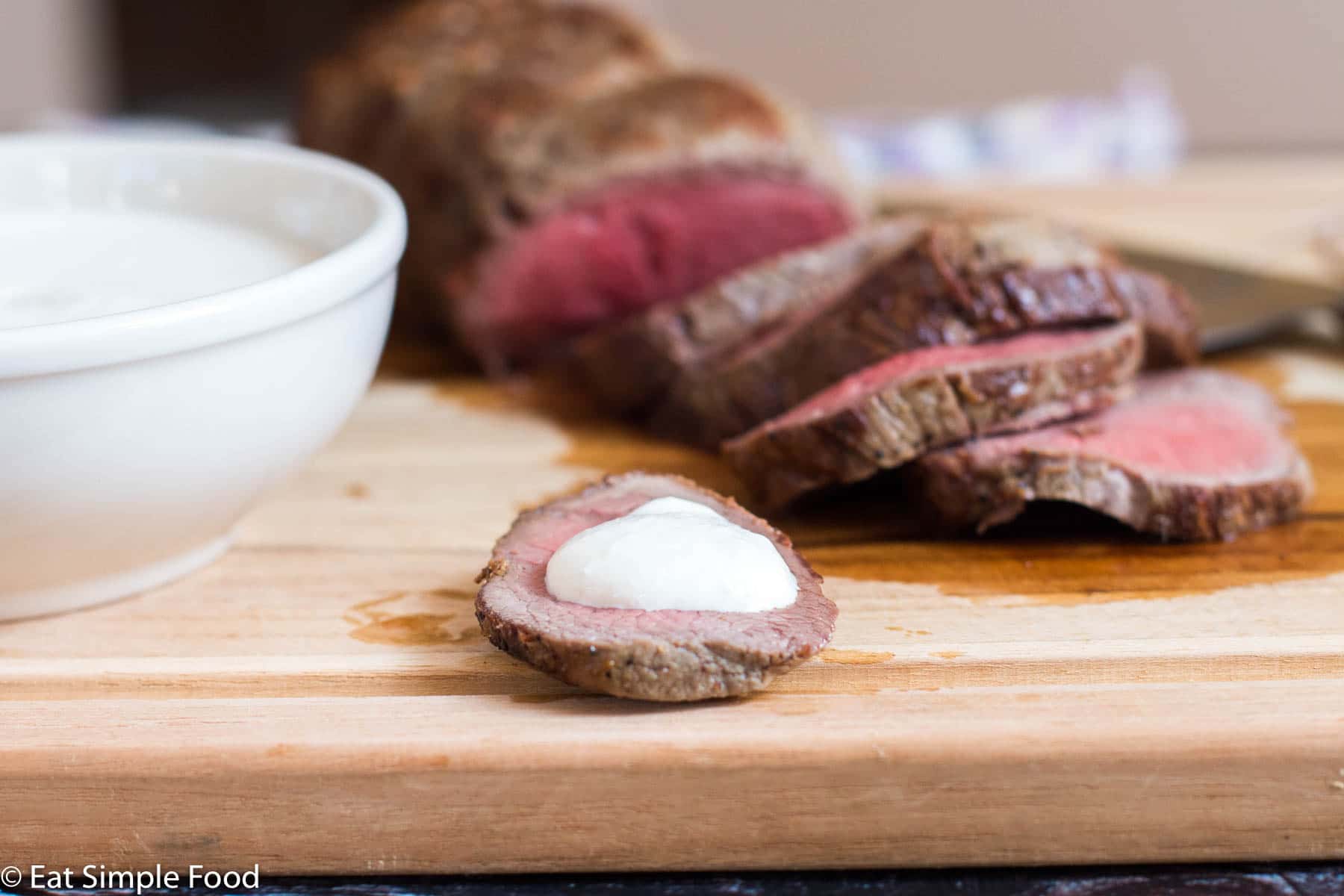 Sliced Rare Beef Tenderloin with One piece with white horseradish sour cream sauce. On a cutting board.