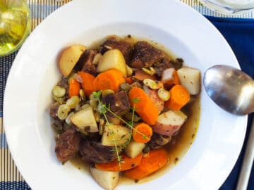 White shallow bowl filled with cooked browned beef, chunks of carrots and potatoes in a beef broth with thyme sprigs on top. Spoon and glass of white wine on side.