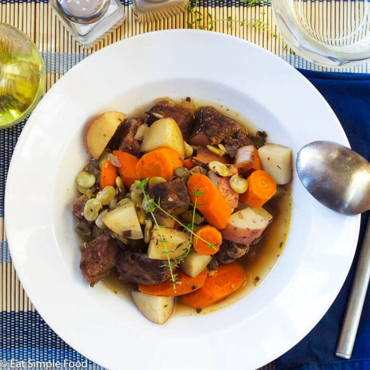 White shallow bowl filled with cooked browned beef, chunks of carrots and potatoes in a beef broth with thyme sprigs on top. Spoon and glass of white wine on side.