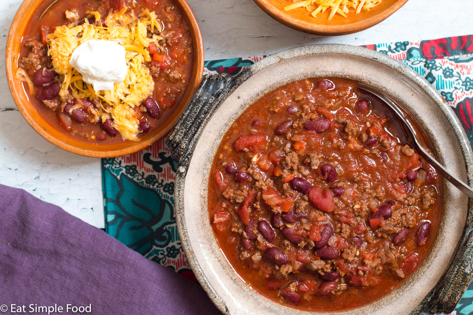 Beef and bean chili in a large brown bowl with a small wood bowl of chili with grated chatter and a spoonful of sour cream.