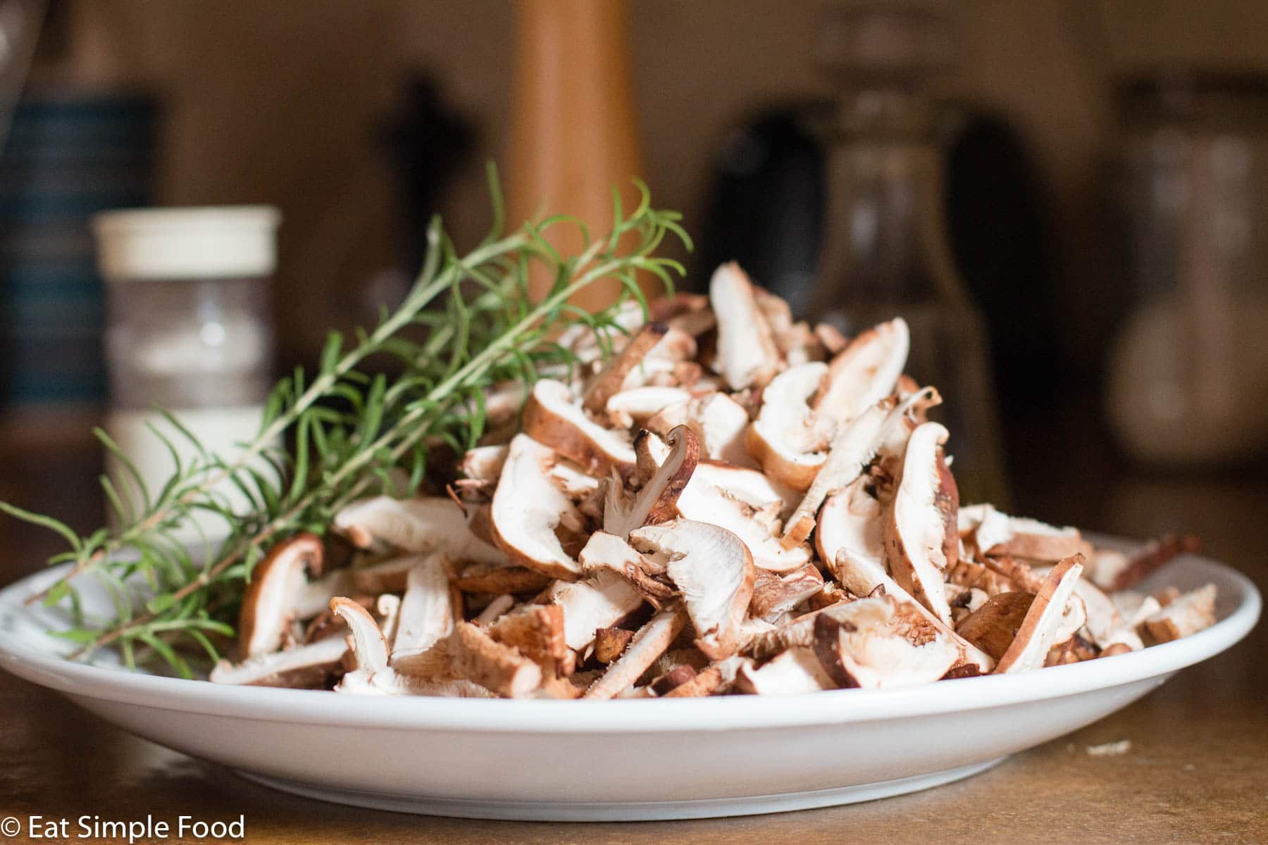 Raw Sliced Shiitake Mushrooms in a white bowl with 4 rosemary sprigs on a brown concrete table.