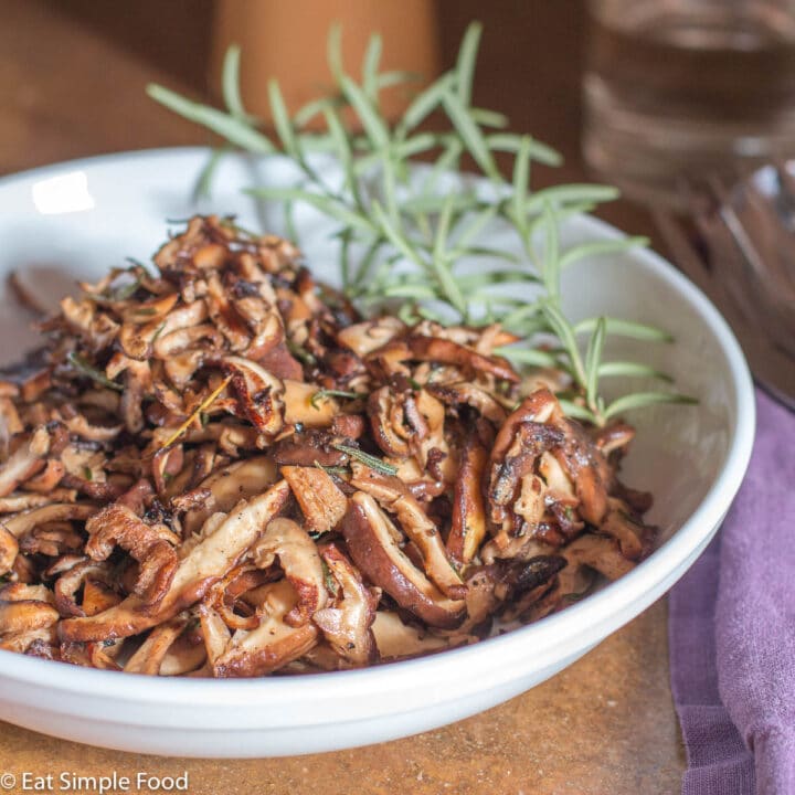 White bowl of crispy shiitake mushrooms with 4 sprigs of rosemary in it and a purple napkin on the side. Side View.