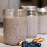 3 Mason jars full of blueberry smoothies. 3 with lids. blueberries and pecans laying on tablecloth in front of them.