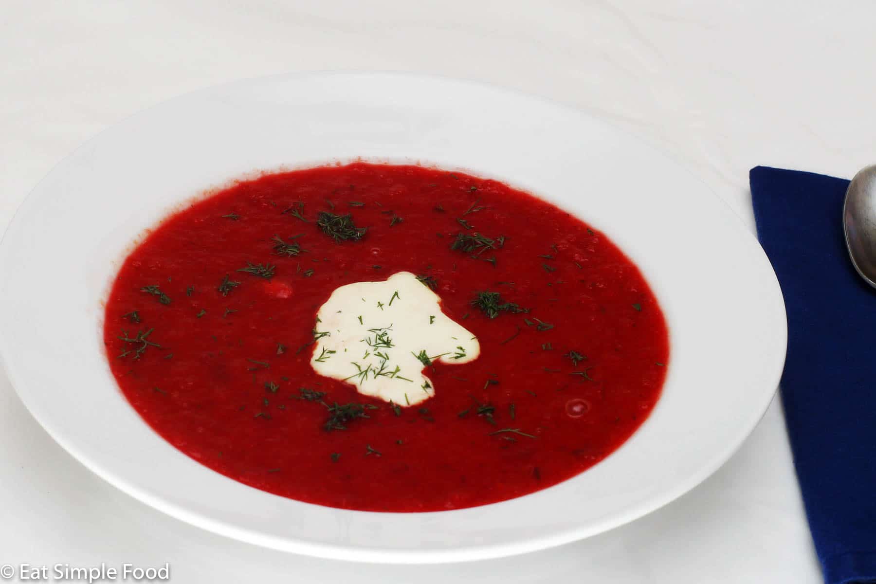 White bowl of vibrant red beet soup (Borscht) with a dollop of sour cream and fresh chopped dill.