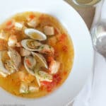 Bouillabaisse (Fish or Seafood Stew) In a white bowl with clams, shrimp, and chunks of white fish in a yellow red delicate sauce.