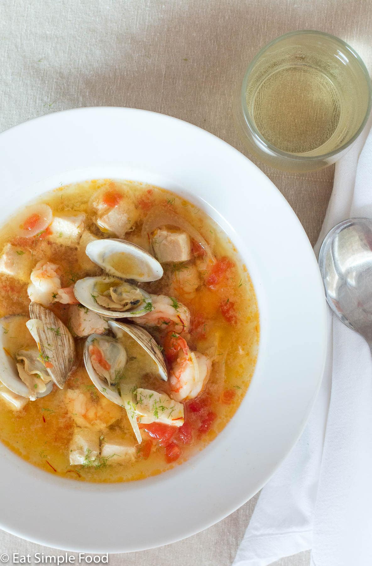 Bouillabaisse (Fish or Seafood Stew) In a white bowl with clams, shrimp, and chunks of white fish in a yellow red delicate sauce.