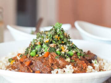 White shallow bowl (side view) of brown/red chunks of lamb with diced carrots and sauce over white rice with a pea, mint, and almond garnish.