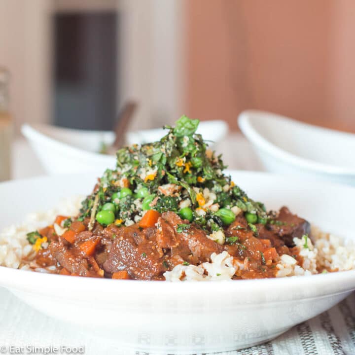 White shallow bowl (side view) of brown/red chunks of lamb with diced carrots and sauce over white rice with a pea, mint, and almond garnish.