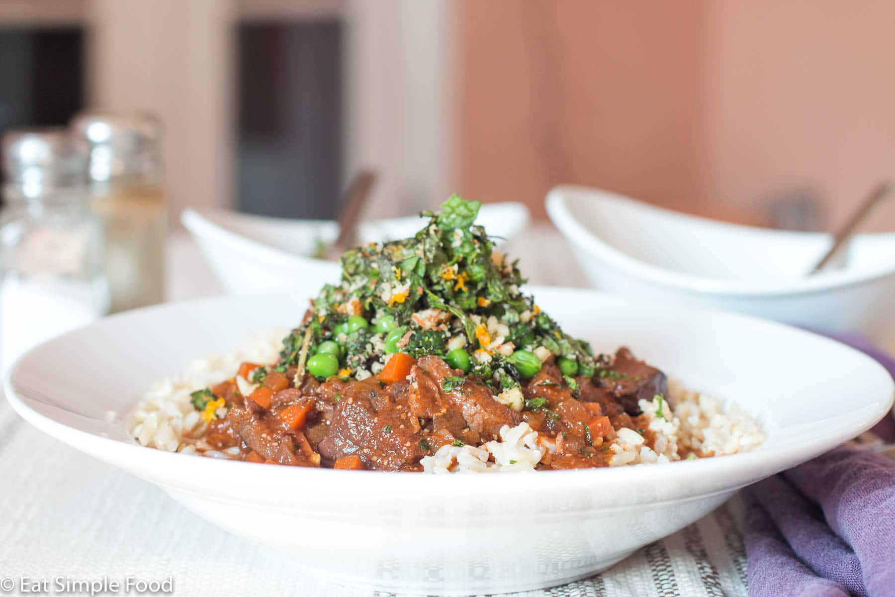 White shallow bowl (side view) of brown/red chunks of lamb with diced carrots and sauce over white rice with a pea, mint, and almond garnish.