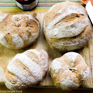 4 loaves of fresh bread on a wood cutting board with jam in the background.