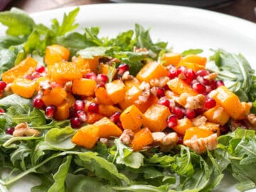 Roasted butternut squash on top of arugula on a white plate. Garnished with chopped walnuts and red pomegranate kernels. side view.