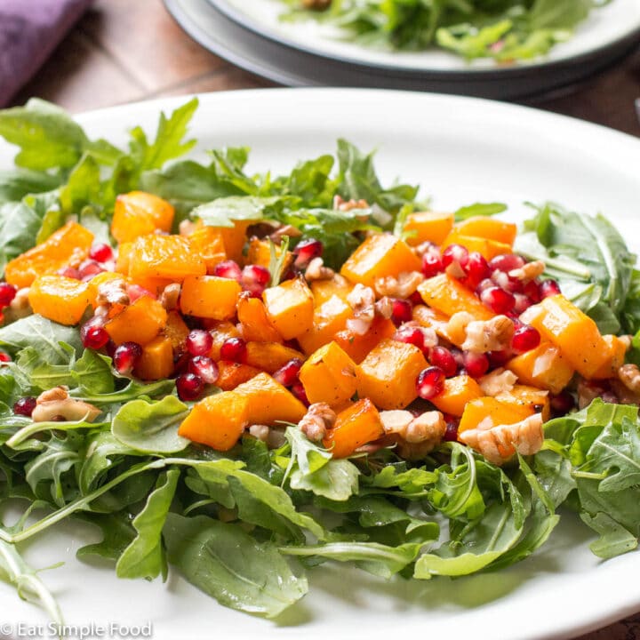 Roasted butternut squash on top of arugula on a white plate. Garnished with chopped walnuts and red pomegranate kernels. side view.