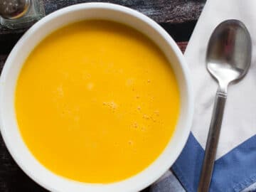 Creamy Orange / Yellow Squash soup in a white bowl. White napkin, spoon and black pepper shaker on the side. Top view.