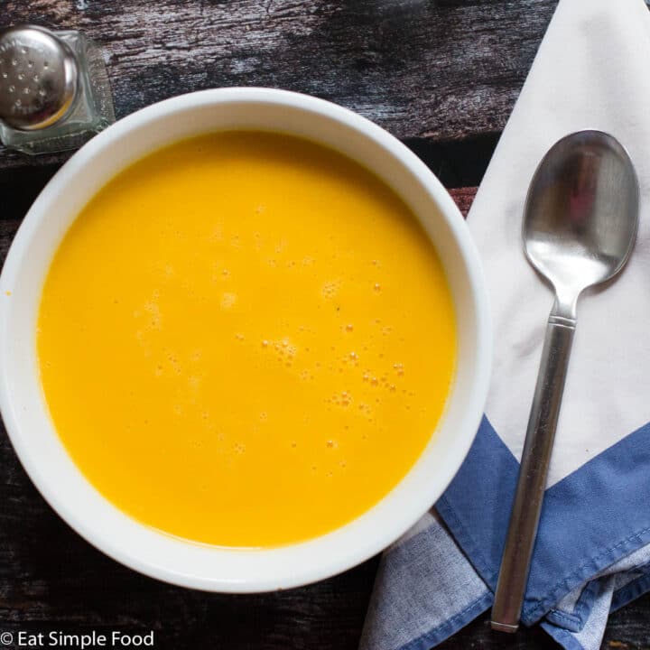 Creamy Orange / Yellow Squash soup in a white bowl. White napkin, spoon and black pepper shaker on the side. Top view.