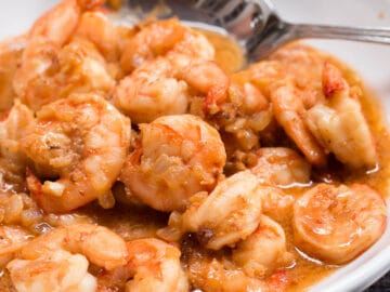 Sauteed Shrimp In A Red/Brown Butter & Worcestershire Sauce In A White Bowl with a Spoon.