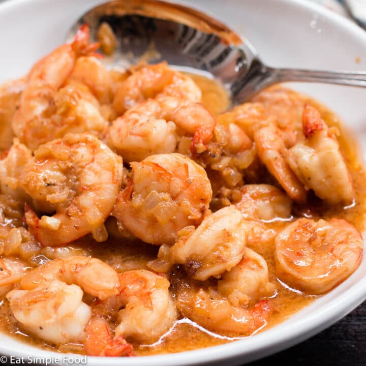 Sauteed Shrimp In A Red/Brown Butter & Worcestershire Sauce In A White Bowl with a Spoon.