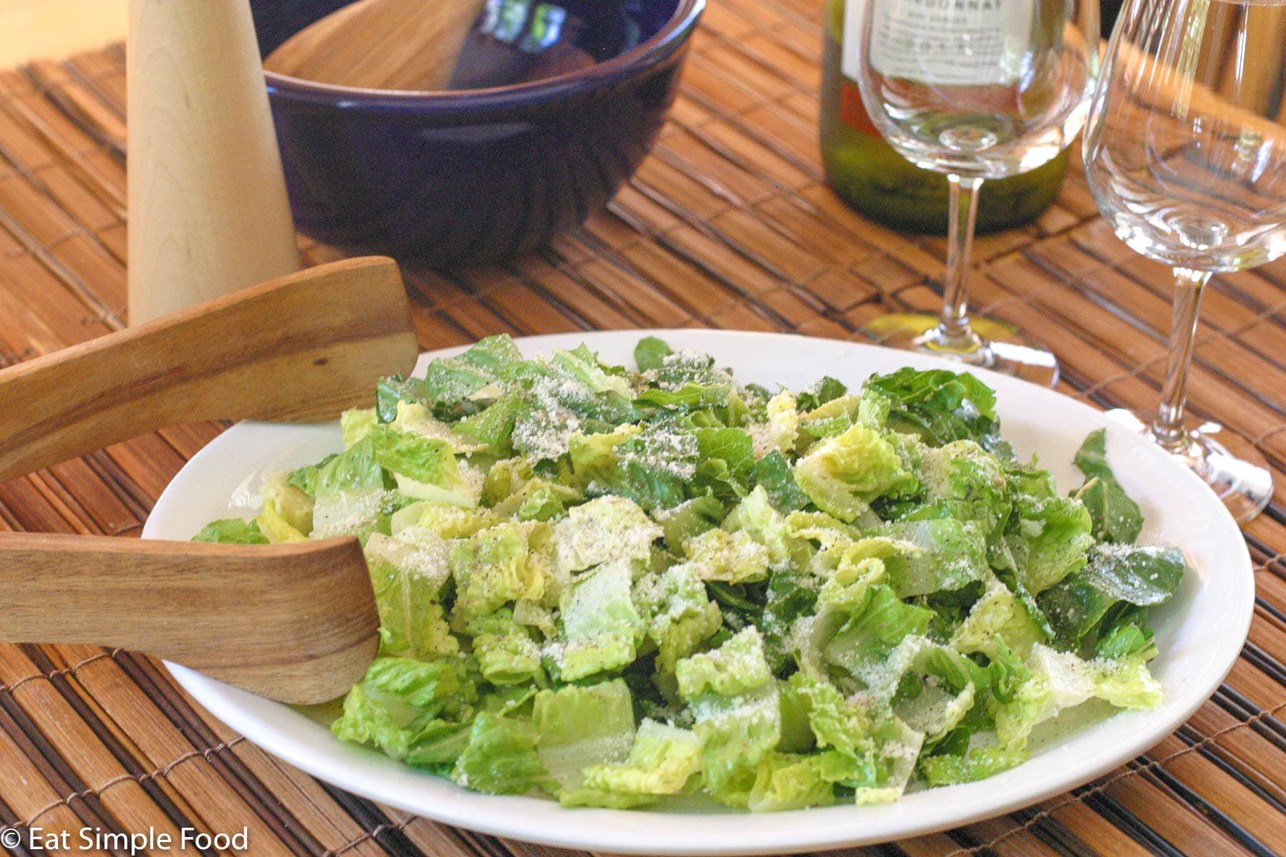 Chunked Romaine Lettuce on a white platter dusted with Parmesan cheese. Tongs on side. 2 glasses of empty wine glasses on side.