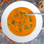 Creamy Orange Carrot Soup in A White Bowl with a brick background with carrots peels around it. Garnished with chopped green cilantro.