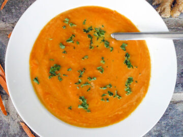 Creamy Orange Carrot Soup in A White Bowl with a brick background with carrots peels around it. Garnished with chopped green cilantro.