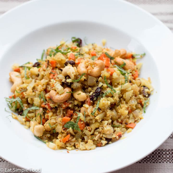 Cauliflower rice with whole cashews, raisins, carrots with curry and garnished with mint. White bowl with spoon. Side view.