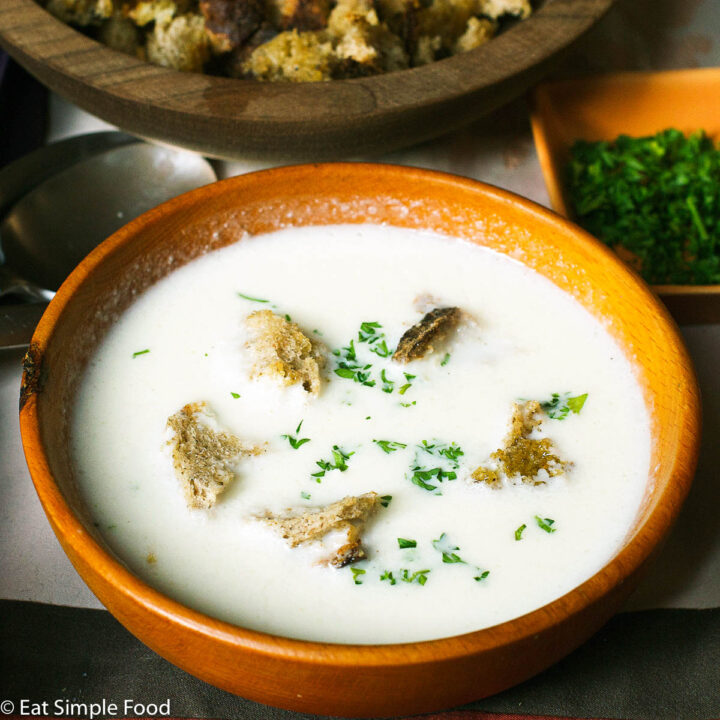 Side View wood bowl of white cauliflower soup with 5 croutons and a chopped parsley garnish