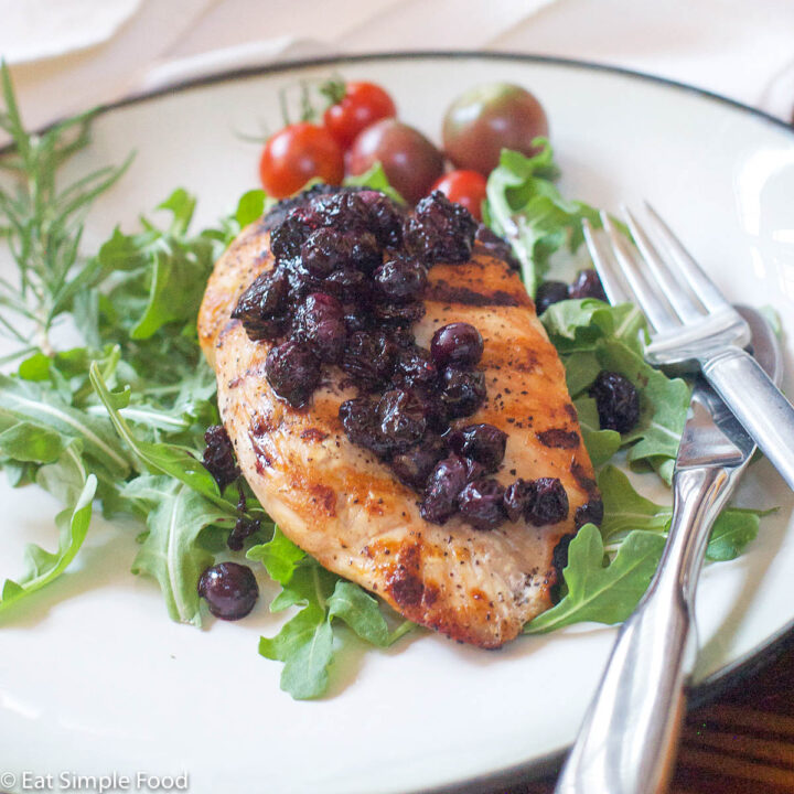 2 white plates with salad with a grilled chicken breast on top with blueberry chutney on top of chicken. Whole Cherry Tomatoes on plates.