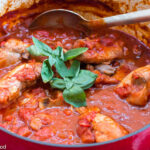 Chicken thighs cooked in a red cast iron pot with a marinara sauce and basil leaf garnish. Serving spoon inside the pot. Side view.