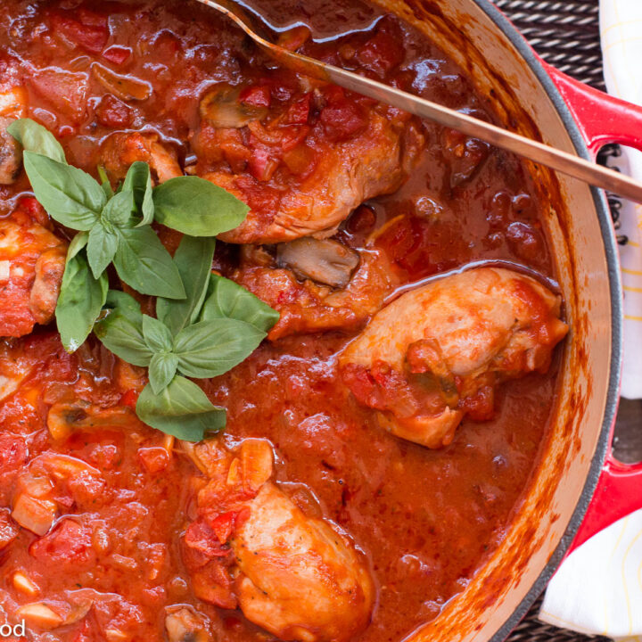 Chicken thighs cooked in a red cast iron pot with a marinara sauce and basil leaf garnish. Serving spoon inside the pot. Top view.